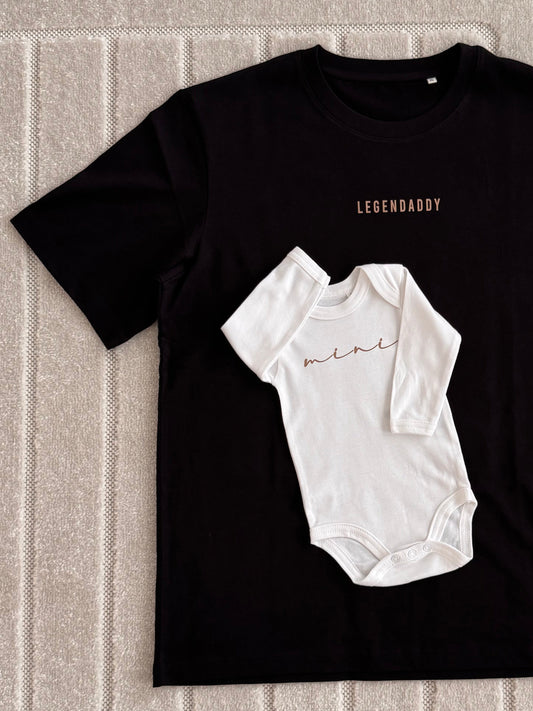 A black short-sleeve t-shirt laid flat on a textured beige carpet, featuring a white baby bodysuit graphic in the center with the word 'mini' in brown script, and the brand name 'LEGENDADDY' printed in brown above the graphic.