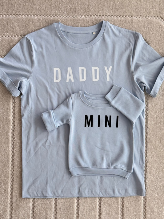 A matching family apparel set featuring a light blue-gray short-sleeve t-shirt with 'DADDY' printed in white across the chest, and a smaller matching short-sleeve t-shirt underneath with 'MINI' printed in black.