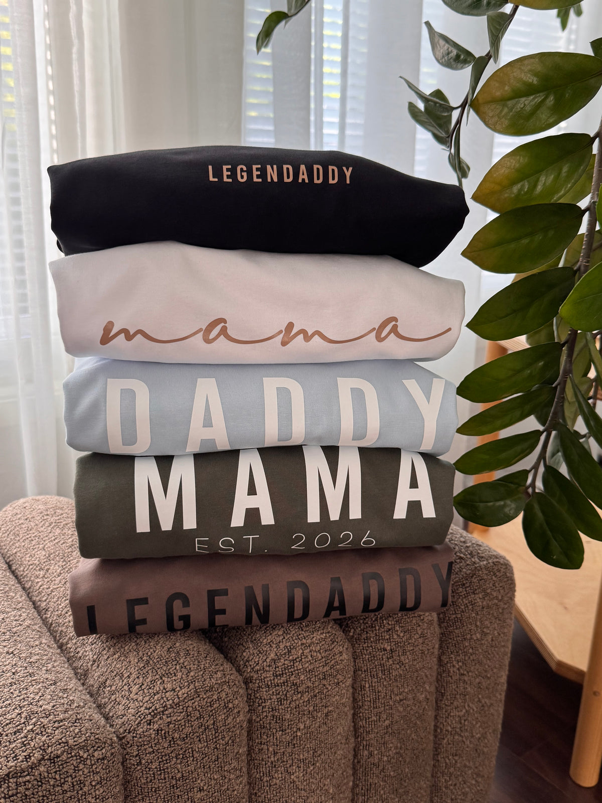 A stack of five folded sweatshirts in different colors sits on a brown upholstered chair in a home setting. The stack includes black, light pink, light blue, dark gray, and brown sweatshirts, each with 'LEGENDDADDY' or 'MAMA' text printed on them.