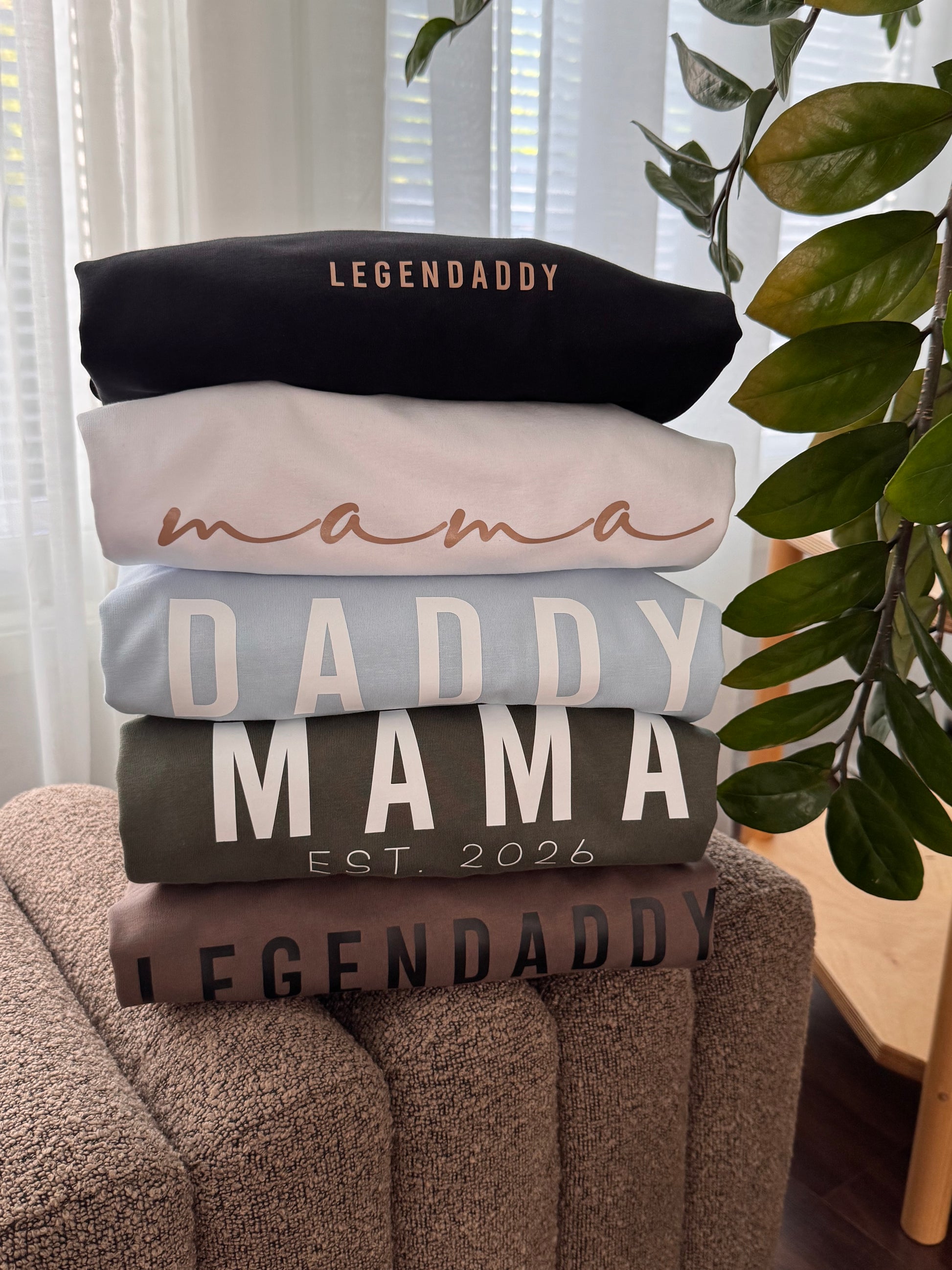 A stack of five folded sweatshirts in different colors sits on a brown upholstered chair in a home setting. The stack includes black, light pink, light blue, dark gray, and brown sweatshirts, each with 'LEGENDDADDY' or 'MAMA' text printed on them.