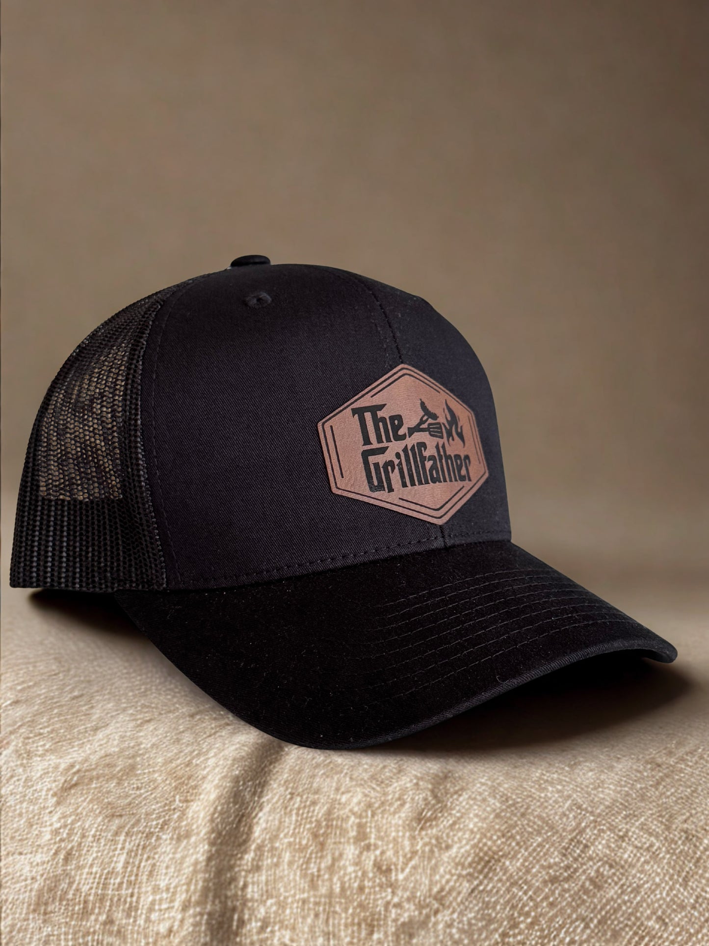 Black trucker cap with 'The Grillfather' logo on a beige surface