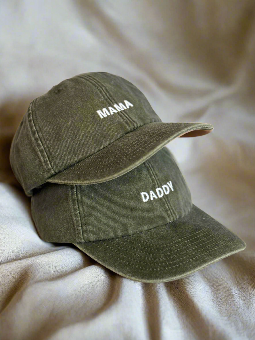 Two olive green baseball caps with 'MAMA' and 'DADDY' text on a beige surface.
